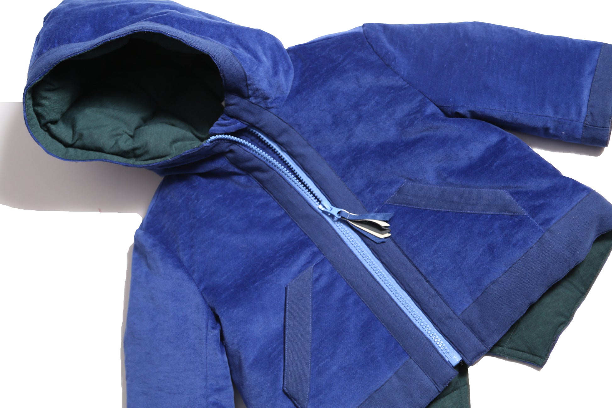 Baby coat made from Blue Breton Tarp Velvet, featuring down filling, an attached hood, slanted ribbon welt pockets, a V cut-out vent at the back hemline, and a center front zipper with a multi-colored ribbon pully for added flair. Lined with soft jersey at the body and nylon lining in the sleeves, by Tia Cibani.