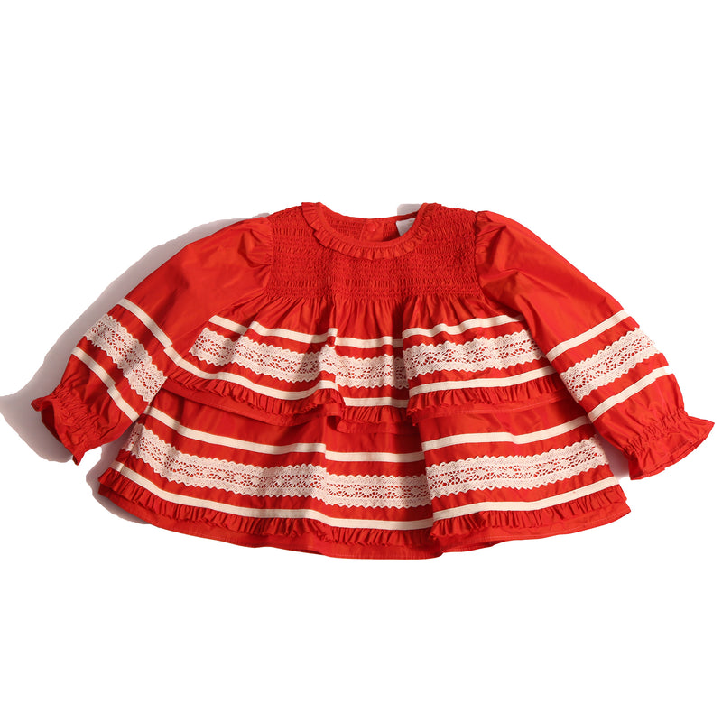 Darling baby smocked blouse in Rouge color, made from luxurious Taffeta with delicate lace embroidered ribbon trim throughout. Features tonal elasticized smocking at the neckline, snaps at the back for easy dressing, and cinched long sleeves with elastic cuffs for a snug fit. Adorned with tiers of small ruffles at each lace appliqué, perfect for special occasions, by Tia Cibani.