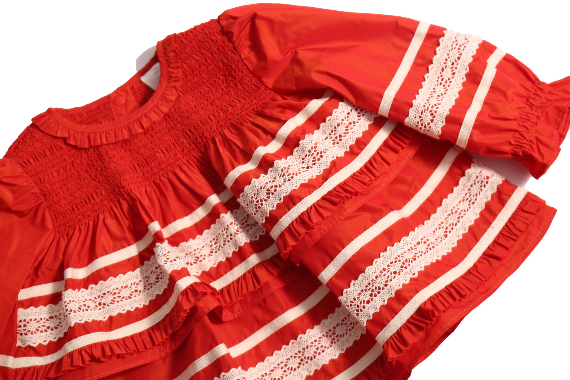 Darling baby smocked blouse in Rouge color, made from luxurious Taffeta with delicate lace embroidered ribbon trim throughout. Features tonal elasticized smocking at the neckline, snaps at the back for easy dressing, and cinched long sleeves with elastic cuffs for a snug fit. Adorned with tiers of small ruffles at each lace appliqué, perfect for special occasions, by Tia Cibani.