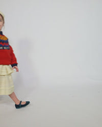 Girl wearing Long-sleeve sweater primarily red, with accents of blue and orange, made of Yarn fabric / Girl wearing a Tealength two-tiered taffeta skirt with elasticized smocking at the waistband, embroidered lace trim, and ribbon at each tier. Unlined for lightweight feel. Colour: Opal, by Tia Cibani. Front View.