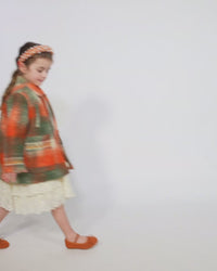 Girl wearing Loden style jacket in cozy Celtic Shadow Plaid, padded and lined for warmth. Snap closure, boxy yet tailored fit with sharp collar, roomy front pockets, and embroidery design at sleeve cuffs. Colour: Multicolour, mainly orange, Tia Cibani.